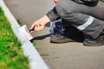 A worker in overalls paints a border with white paint on a summer day. Urban services, landscaping. Hand holds a paint brush. Close-up