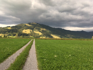 View of straight country road through pasture and stormy sky above alpine mountain