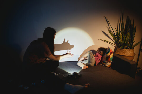 mother and child play at home on sofa with lantern in shadow on wall. mom shows her daughter dog or wolf out of her hands. kids games and fun