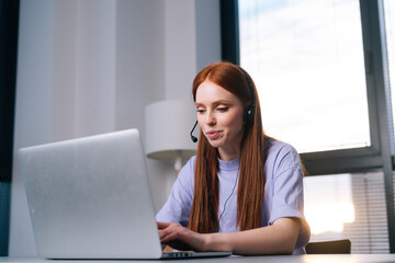 Close-up of professional young woman operator using headset and laptop during customer support at home office. Young redhead female student communicating online by video call on background of window.