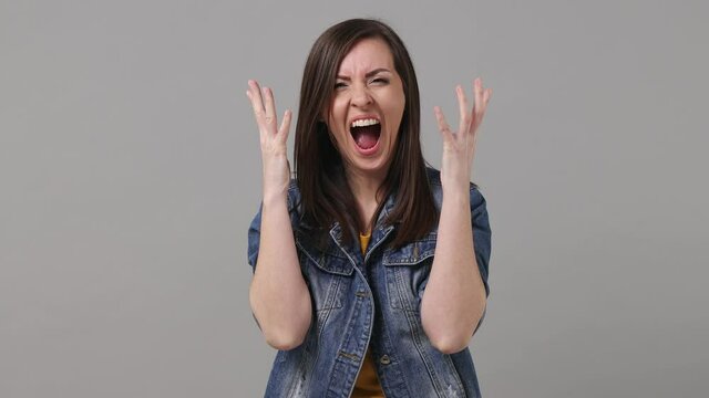 Displeased irritated sad angry mad brunette woman 20s years old in denim jacket yellow t-shirt scream swear shout call out expressive gesticulating with hands isolated on grey color background studio