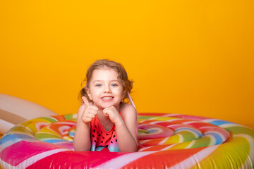Fototapeta na wymiar Happy little girl in swimming suit lying on colorful inflatable mattress lollipop on yellow background.