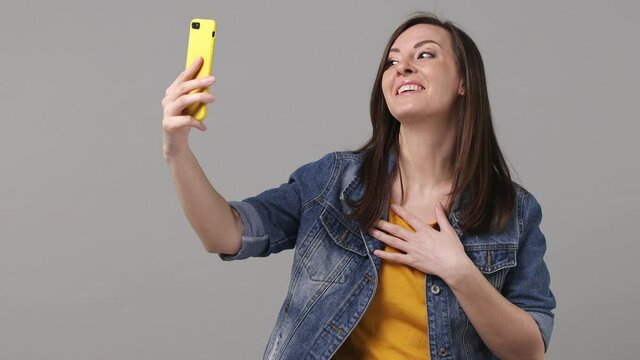 Young woman 20s years old in denim jacket yellow t-shirt get video call mobile cell phone doing selfie talk conducting pleasant conversation greet with hand isolated on grey color background studio