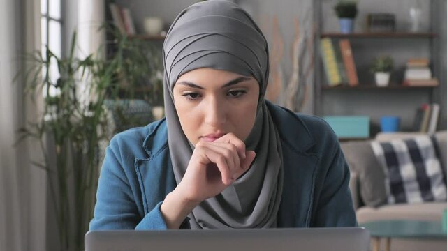 pensive arab muslim woman wearing hijab having a work problem at the laptop thinking of a solution,concerned female middle eastern in headscarf in front of computer has trouble looking for inspiration