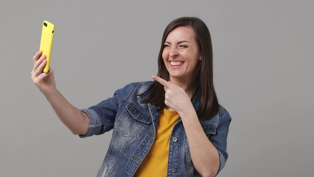 Charming fun beautiful brunette young blogger woman 20s years old in denim jacket yellow t-shirt doing selfie shot on mobile phone post photo on social network isolated on grey color background studio
