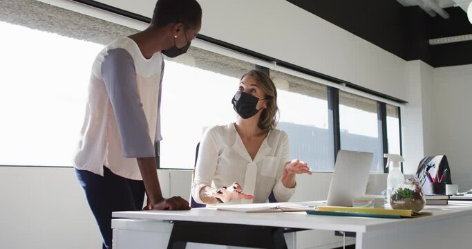 Two diverse female colleagues wearing face masks looking at laptop and discussing in office
