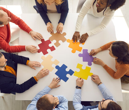 Team of young and senior employees sitting in circle around office table putting jigsaw puzzle together as metaphor for teamwork and looking for working business solutions. Square format, high angle - Powered by Adobe