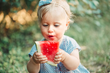  Summer seasonal picnic food. Cute baby girl eating ripe red watermelon in park. Funny child kid...