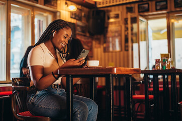 Cheerful african american woman using smartphone and airpods while sitting in a cafe