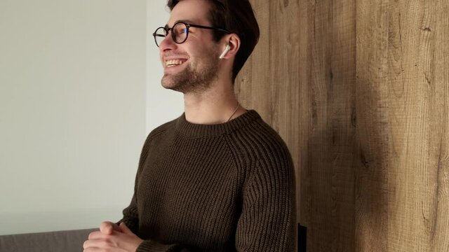 A smiling man in a sweater talking on wireless headphones and actively gesticulating while walking in the room