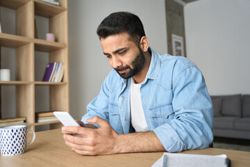 Young Indian Hispanic businessman using smartphone working remotely reading ebook from modern home office. Latin student having virtual training on cellphone using online tech applications.