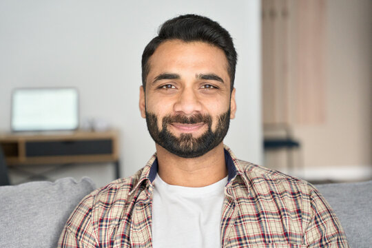 Headshot Portrait Of Attractive Confident Indian Hispanic Man Looking At Camera Sitting On Coach At Modern Living Room. Latin Businessman Posing In Casual Stylish Look At Home Office.