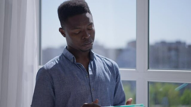Portrait of African American man using tablet thinking standing at window in home office indoors. Intelligent smart young freelancer analyzing e-business planning startup idea. Creative thinking
