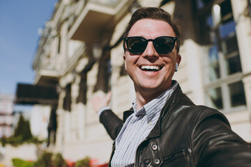 Close up young smiling fun caucasian man 20s wearing black leather jacket glasses doing selfie shot on mobile cell phone camera show downtown buildings, Concept of people urban lifestyle excursion