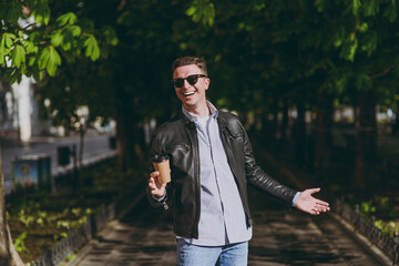 Young european happy friendly man in black leather jacket eyeglasses strolling in green park hold takeaway delivery craft paper cup drink coffee to go Concept of people urban lifestyle spring weather