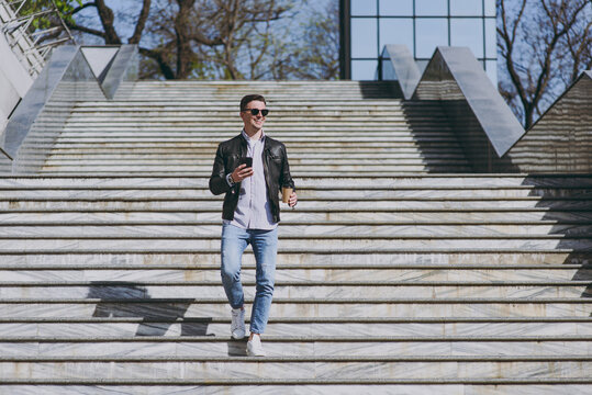 Full length smiling young man in leather jacket eyeglasses walk in city downtown go down concrete steps hold mobile cell phone, takeaway craft paper cup drink coffee to go Concept of urban lifestyle.