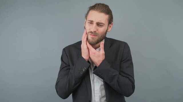 A young long-haired businessman suffers from dental pain keeping his pike. Hipster man isolated in studio on gray background