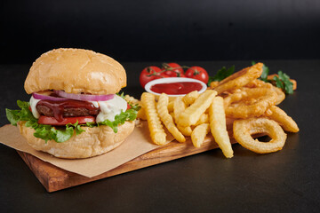 Homemade hamburger or burger with fresh vegetables and cheese lettuce and mayonnaise served, French fries on pieces of brown paper on black stone table . concept of fast food and junk food