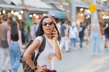 Happy young travel asian woman using mobile phone and relax on street chatuchak weekend market in bangkok, holiday with bags, sunglasses,Thailand, Travel vacation city concept.