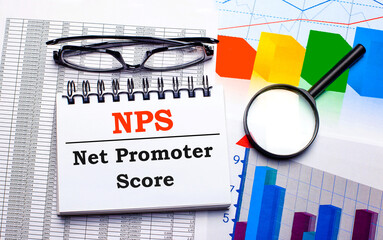 On the desktop are glasses, a magnifying glass, color charts and a white notebook with the text NPS Net Promoter Score. Business concept. View from above