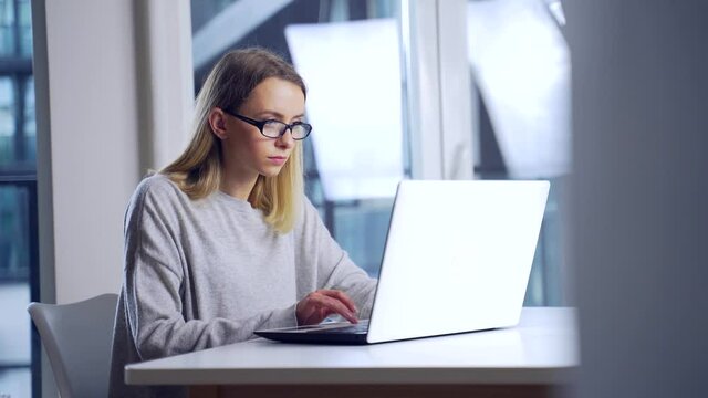 young female student working in the evening on a laptop at workplace herself. Woman freelancer alone in office or home typing front computer screen in modern room. Online learning or distance work