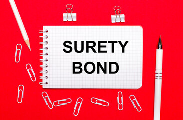 On a red background, a white pen, white paper clips, a white pencil and a notebook with the text SURETY BOND. View from above