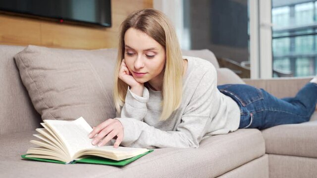 young woman sitting or lying on sofa at home and reading a book. student girl studying lessons. female relaxes indoors on a comfortable couch at leisure. nice to spend free time on the weekend