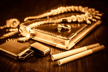 Golden lighter with a leather diary and cigarettes with jewellery on a mahogany table. Focus on the...