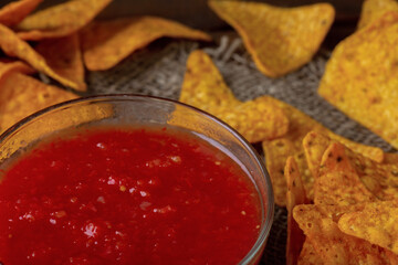 Closeup corn chips and tomato sauce in glass bowl on burlap background. Nachos with red sauce on dark table. Traditional mexican tortilla snacks
