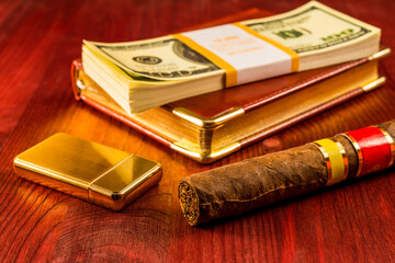 Pack of dollars and golden lighter with a leather diary and cuban cigar on a mahogany table. Focus...