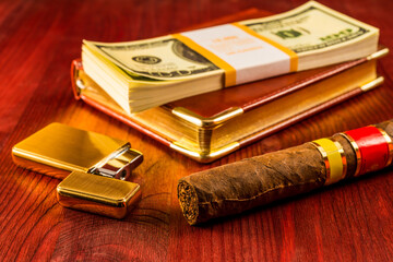Pack of dollars and golden lighter with a leather diary and cuban cigar on a mahogany table. Focus on the cuban cigar