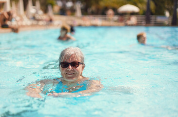 Elderly senior woman with grey hair, wearing blue swimsuit smiling in hotel pool