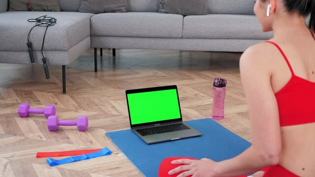 Fit woman in sportswear sits on yoga mat looks computer display, tells listen fitness trainer online video call webcam chat, distance sport course. Green screen mock up chroma key monitor laptop