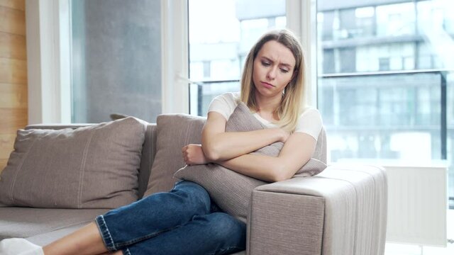 Portrait young woman sitting on sofa at home depressed. Girl is having a hard time with stress. Caucasian female indoors alone worried about problems. unhappy blonde student in living room or bedroom