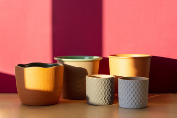 five empty ceramic pots in terracotta, brown, white and gray shades for planting indoor plants against a maroon wall on a wooden table in bright sunlight with hard shadows