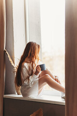 A young girl with long hair sits on a window sill with a cup in her hands