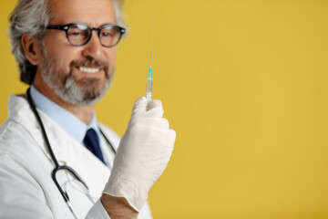 Mature doctor in white medical coat and white gloves posing with syringe against yellow background.