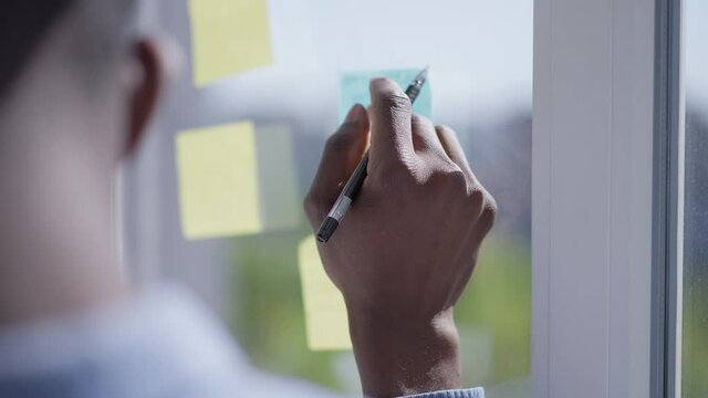 Close-up of African American young male hand writing with pen on sticky notes on window. Unrecognizable man handwriting business ideas in home office indoors on sunny day