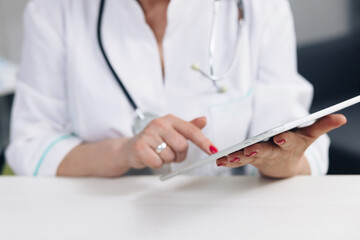 Close up middle aged older female doctor in white uniform holding digital computer tablet in hands, managing patients visits. Mature physician checking health history data in gadget, using modern app