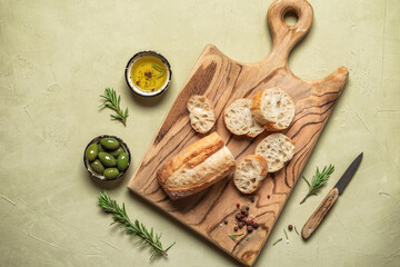 Fresh ciabatta slices on a wooden board with olive oil, olives and rosemary. Beige concrete background. Italian bread. Top view, flat lay.