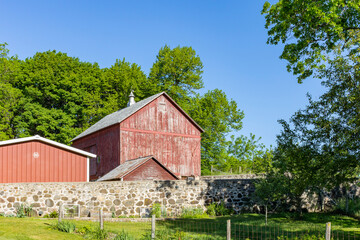 Fototapeta na wymiar A small faded red barn with a milk house, shed, and stone barnyard wall on a clear sunny day.