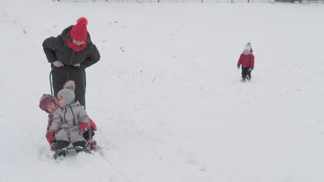 vacation, game, family concept - slo-mo authentic three happy preschool toddler kids siblings with young wonan mom sledding and play with snow. snowfall in cold season weather in winter yard outdoors