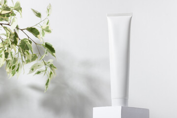 White cosmetic tube on a stand with leaves and shadows. Toothpaste, face and body cream. Female cosmetic tube with skin care product. Organic cosmetics. Copy space.