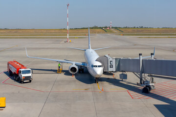 White airplane docked on apron outside airport terminal with jet bridge. Worker refuels plane. Fuel...