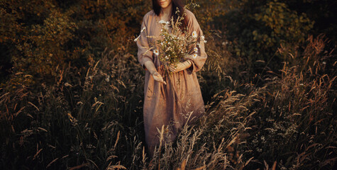 Beautiful woman in linen dress gathering wildflowers in summer meadow in evening. Stylish young...