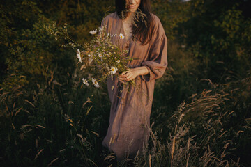 Beautiful woman in linen dress gathering wildflowers in summer meadow in sunny evening. Stylish...