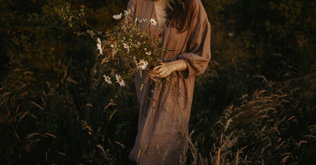 Beautiful woman in linen dress gathering wildflowers in summer meadow in evening. Atmospheric...