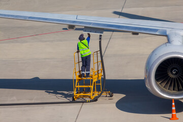 Rear view of male worker in green vest refueling airplane while standing on ladder on airfield....