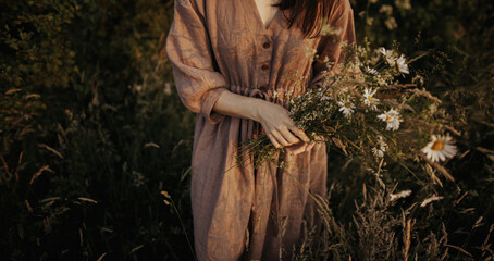 Beautiful woman in linen dress gathering wildflowers in summer meadow in evening.  Stylish young female in rustic dress picking flowers in countryside. Atmospheric stylish vintage image