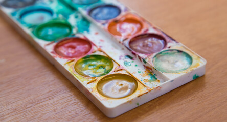 On the table is a used set of multicolored watercolor paints for painting. Top view of half-empty jars of watercolors. Flat layout on the table.